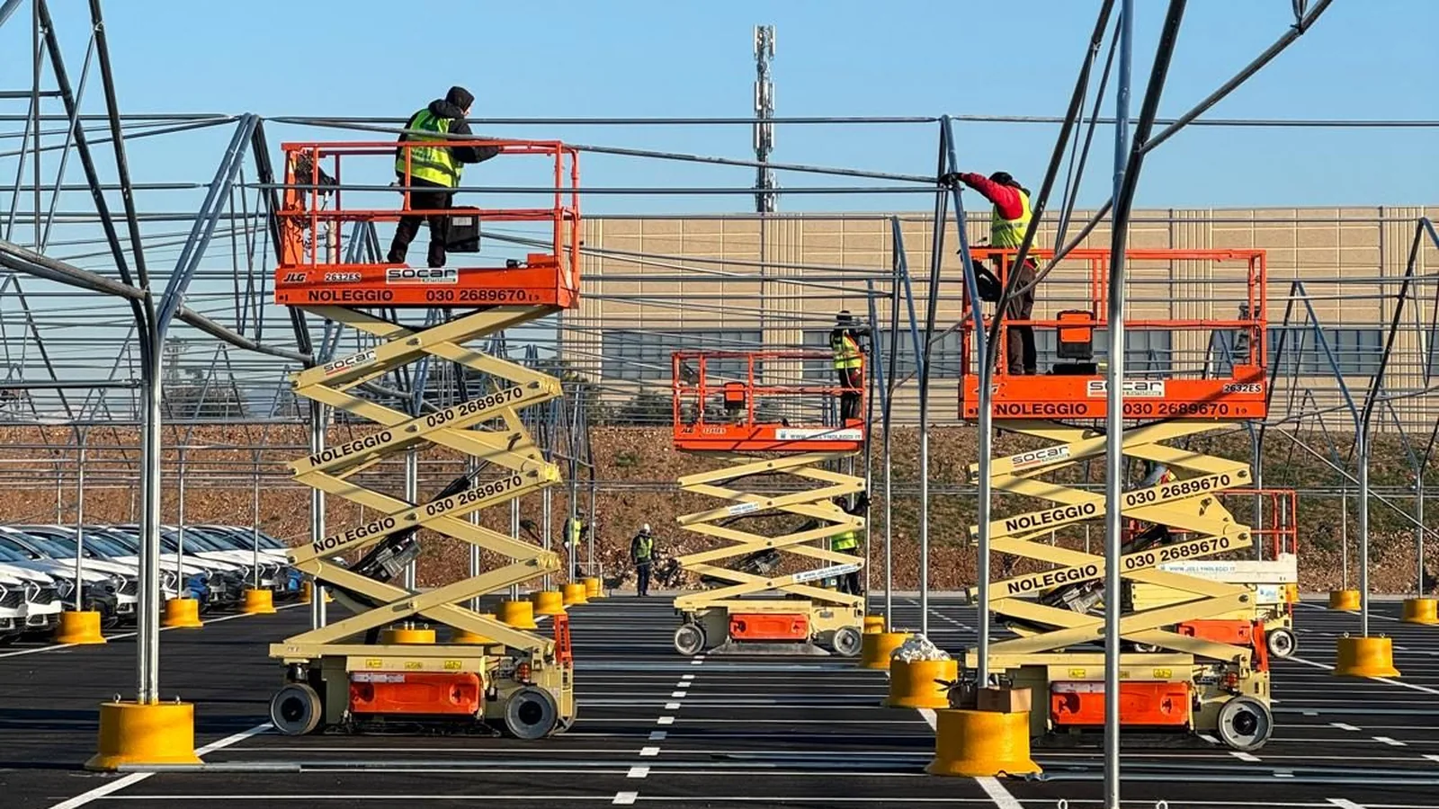 Workers on scissor lifts assembling a Euromet modular cover structure over a large logistics yard with parked vehicles