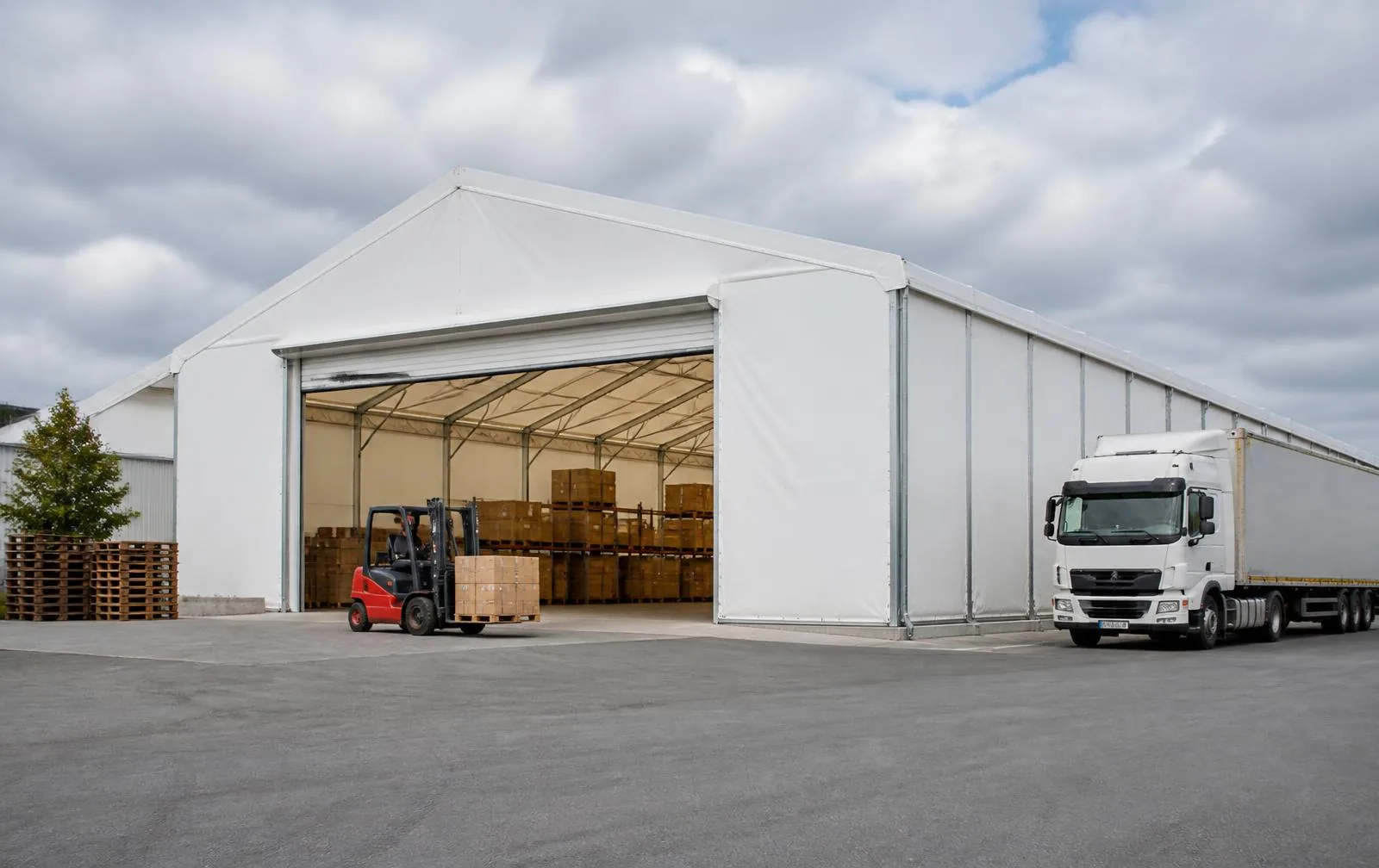 White PVC industrial hangar with galvanised steel frame in a logistics yard, open door with a forklift and a truck manoeuvring outside